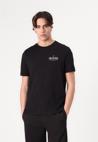 Zwarte katoenen t-shirt met ronde hals, korte mouwen en een klein wit "HILFIGER" logo op de linker borst. Glad textuur.