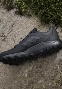 Scarpa sportiva nera con suola robusta appoggiata sul terreno accanto all'erba.