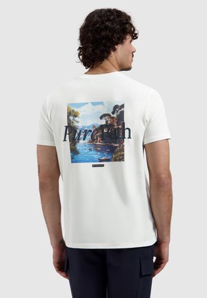 Man met krullend haar, gekleed in een wit T-shirt met een kleurrijk kustlandschap en de tekst "Pure Earth" op de achterkant, staand tegen een effen achtergrond.