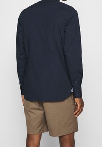 Maglietta blu a maniche lunghe con polsini con bottoni, colletto piatto e taglio dritto, indossata sopra pantaloni beige. Tessuto liscio con dettagli minimi.