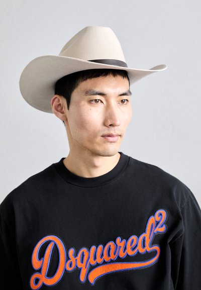 Jovem usando um chapéu de cowboy branco e um moletom preto com grande texto laranja e azul "Dsquared²", olhando ligeiramente para a direita.