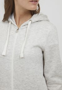 Helle graue Zip-Hoodie aus weichem Stoff, mit Kapuze, weißen Zugbändern und silberfarbener Hardware am Reißverschluss.