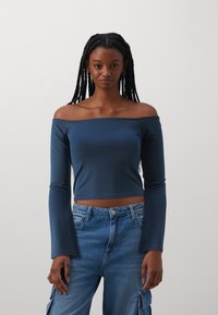 Plava off-the-shoulder dugorukava crop majica od rastezljivog materijala, u kombinaciji s trapericama širokih nogavica s viskim strukom i džepovima.