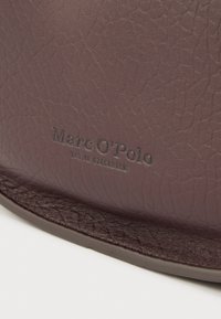 Mørk bordeaux lædertaske med en tekstureret overflade. Har diskret prægning af mærket "Marc O'Polo" langs den nederste kant.