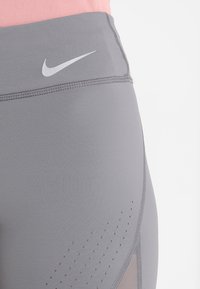 Graue Sportleggings mit hohem Bund, versehen mit einem kleinen weißen Nike-Logo und Mesh-Einsätzen an der Seite für Atmungsaktivität.