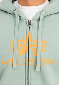 Mentazöld, húzózsinórral ellátott zipzáros kapucnis pulóver, elöl narancssárga logóval és "ALPHA INDUSTRIES" felirattal. Ezüst színű zipzár hardver látható.
