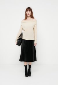 Pulôver bege às costas, combinado com uma saia plissada preta, saltos pretos e meias pretas. Design simples com texturas suaves.