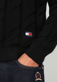 Schwarzer Kabelstrickpullover mit gerippten Bündchen und Saum, ausgestattet mit einem kleinen roten, weißen und blauen Logolabel. Getragen mit dunklen Jeans.