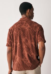 Chemise à manches courtes de couleur rouille, avec un motif texturé matelassé représentant des soleils et des feuilles. Dotée d'un col classique et d'une coupe décontractée.