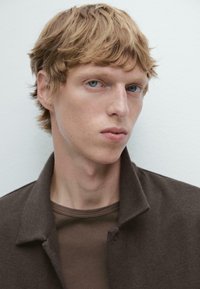 Bruine colbert over een bruine shirt, gecropte close-up van een persoon met kort, golvend blond haar en lichtblauwe ogen tegen een lichte achtergrond.