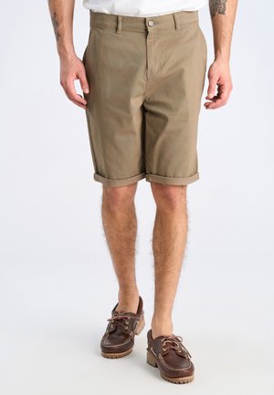 Man wearing rolled-up khaki shorts and brown leather boat shoes, standing against a plain light background.