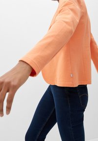 Top de manga larga naranja con cuello redondo y dobladillo curvado, combinado con vaqueros ajustados oscuros. La tela parece suave con una textura sutil.