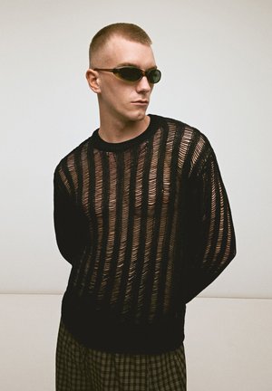 Junger Mann mit kurzen Haaren, der schwarze getönte Sonnenbrillen, einen schwarzen transparent gestreiften Pullover und karierte Hosen trägt, steht vor einem schlichten Hintergrund.