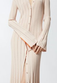 Haljina od svijetlo breskvastog pletiva s dugim rukavima, vertikalnim prugama i kopčanjem na gumbe; ima razdvojen rub i kontrastni narančasti detalj na krajevima rukava.