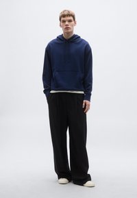 Hanorac bleumarin cu glugă și buzunar frontal, asortat cu pantaloni negri cu picior larg și pantofi bej slip-on, pe un fundal neutru.