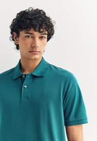 Žalsvai mėlynas polo marškinėlis iš tekstūruotos medžiagos, su apykakle ir trim mygtukais. Kairėje krūtinės pusėje išsiuvinėtas logotipas.