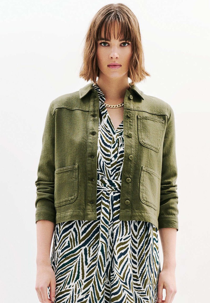 Veste cropped vert olive, tissu texturé, fermeture à bouton, deux poches avant. Portée par-dessus une robe à motifs avec de larges rayures vertes et blanches.