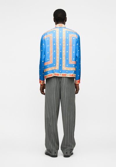 Persoană purtând o cămașă albastră cu mânecă lungă cu model și detalii portocalii și aurii, combinată cu pantaloni largi cu dungi verticale alb-negru.