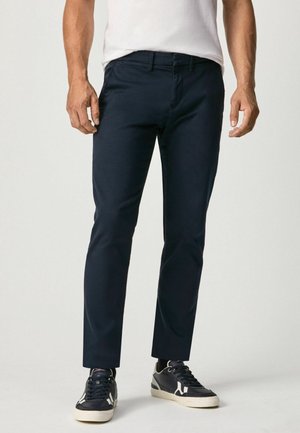 Man wearing slim-fit dark blue pants paired with black and white sneakers, standing against a plain light background.
