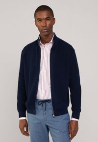 Granatowy kardigan na zamek błyskawiczny z prążkowaną fakturą, wysokim kołnierzem i srebrnymi dodatkami; noszony na jasnej, w paski koszuli o luźnym kroju.