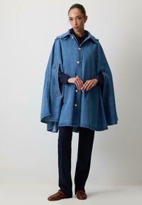 Denimcape i lyseblå, med krave, guldknapper og sidelufter. Båret over sorte bukser og brune loafers.