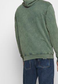 Sweat à capuche vert avec une finition douce et délavée, doté d'une capuche à cordon et de poignets côtelés. Assorti à un jean en denim bleu.