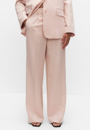 Kvinde iført lyserøde, vidde bukser og matchende blazer, med den ene hånd i lommen og sorte sandaler med firkantet tå synlige.