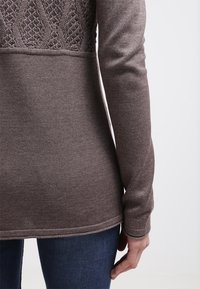 Gros plan sur le dos d’une personne portant un pull en maille marron avec un dos supérieur texturé et un jean bleu foncé, debout avec une main détendue.