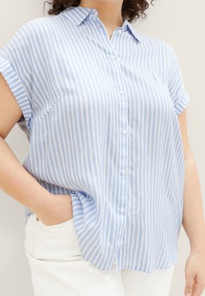 Kobieta ubrana w koszulę na guziki z krótkim rękawem w pionowe paski w kolorze jasnoniebieskim i białym oraz białe spodnie, jedna ręka w kieszeni.