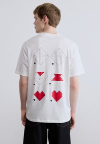 Λευκό βαμβακερό μπλουζάκι με μαύρες και κόκκινες γραφικές απεικονίσεις στην πλάτη, που απεικονίζουν γεωμετρικά σχήματα και σχέδια καρδιάς.