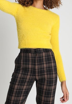 Nainen, joka pukeutunut kirkkaan keltaiseen pörröiseen pitkähihaiseen lyhytmalliseen villapaitaan ja korkeavyötäröisiin mustiin ruutuhousuihin, joissa on ruskeita viivoja.