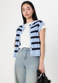Hellblauer, gestrickter Cardigan mit marineblauen Streifen, runder Halsausschnitt, kurzen Ärmeln und goldenen Knöpfen; kombiniert mit einem weißen Hemd und Jeans.