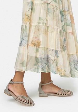 Pies con sandalias planas plateadas y brillantes con tiras en el tobillo, combinadas con una falda midi fluida de estampado floral claro.