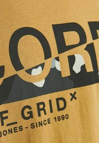 Żółta bawełniana koszulka z odważnym czarnym napisem pod spodem z kamuflażowym wzorem. Zawiera logo z napisem "OFF GRID" oraz "JONES - SINCE 1990."
