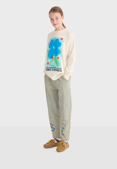 Young person in beige long-sleeve shirt with large blue flower graphic, relaxed fit light pants with blue patterns, and brown moccasin shoes.