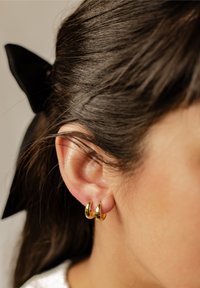 Boucles d'oreilles en or avec une surface polie, accrochées à une oreille. La coiffure est lisse avec un accent de nœud noir. Un éclat subtil met en valeur leur design.