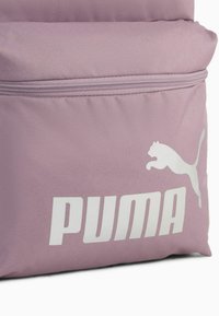 Mochila de tela morada con un acabado texturizado, que presenta un logo blanco de "PUMA" y un gráfico de un gato saltando. Compartimento con cremallera en la parte frontal.