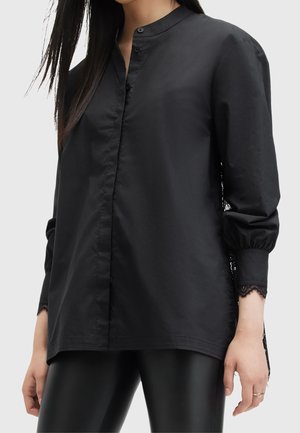 Femme portant une chemise noire ample boutonnée avec un bord en dentelle au poignet et un pantalon en cuir noir sur un fond uni.