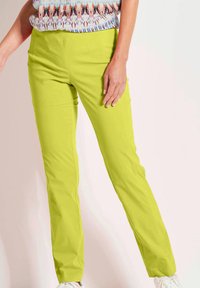 Limegrüne, enganliegende Hose mit glatter Textur, ausgestattet mit Fronttaschen und einem eleganten Design. Kombiniert mit einem gemusterten Oberteil und weißen Sneakers.