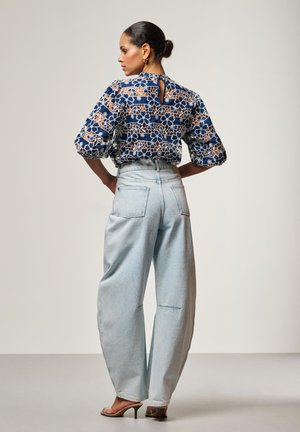 Vrouw met haar in een knot, gekleed in een marineblauwe blouse met bloemenkant en lichtblauwe wijde jeans met hoge taille, gecombineerd met naakte sandalen met hak, poseert met handen in de zij.