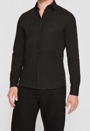 Man draagt een nauwsluitend, zwart overhemd met lange mouwen en zwarte broek tegen een effen lichte achtergrond.