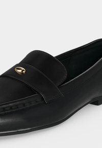 Mocassin en cuir noir à petit talon, avec un détail ovale doré décoratif sur la bride supérieure.