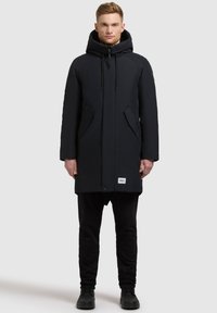 Parka lungă neagră cu glugă, fermoar frontal și sfori reglabile. Dispune de buzunare laterale și un patch cu logo aproape de tiv. Design călduros și căptușit.