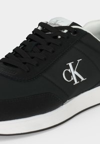 Baskets Calvin Klein noires avec semelle blanche, lacets noirs et logo cK blanc brodé sur le côté.