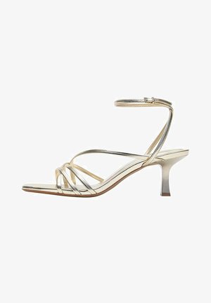 Zlatna sandala s otvorenim prstima, remenčićem oko članka i niskom petom, prikazana na bijeloj pozadini.