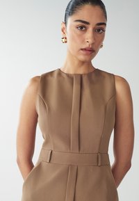 Hnědress bez rukávů s přiléhavým živůtkem, sklady v přední části a staženým pasem. Hladká, strukturovaná tkanina bez viditelných vzorů.