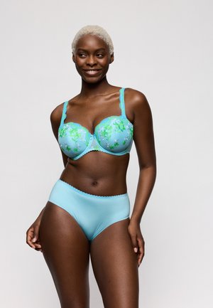 Lachende vrouw met kort blond haar, gekleed in een blauw bloemenkantbeha en bijpassende effen blauwe hoge taille slip, staand tegen een effen achtergrond.