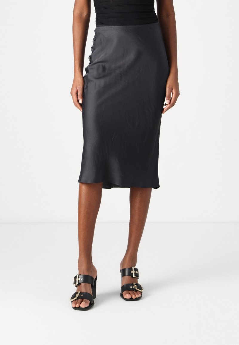 Femme portant une jupe noire midi coupe slip et des sandales noires à grandes boucles dorées, debout devant un fond blanc uni.