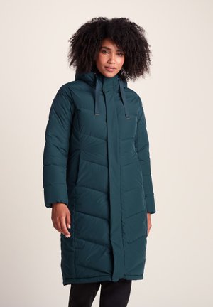 Woman wearing a long dark teal padded winter coat with a hood and drawstrings, standing against a plain light background.