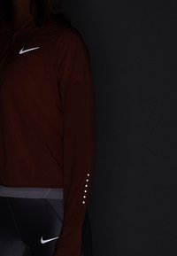 Persona con chaquetaNike de color rojo oscuro y leggings Nike negros, de pie frente a un fondo negro, con los logotipos de Nike visibles en la ropa.